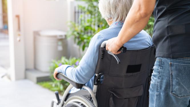 Attēlā vīrietis stumj invalīdu ratīņus kurā sēž sirma kundze