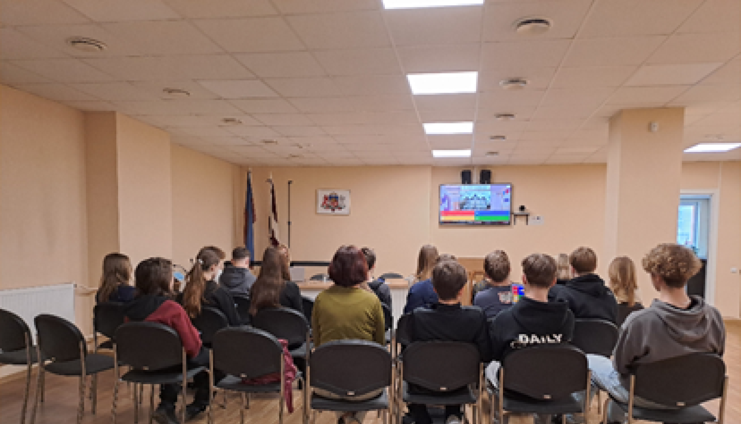 Attēlā attļeoti skolēni, kas seiž telpā uz krēsliem.